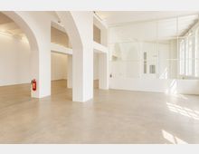 Heller, leerer Innenraum mit weißen Säulen und Rundbögen, glattem Betonboden und Deckenleuchten. Rechts trennt eine große Glaswand mit Sprossen einen Bereich ab; daneben fallen durch hohe Fensterfronten Tageslicht und Schatten auf den Boden.