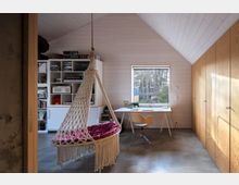 Ein helles Zimmer mit Holzwänden und einem schrägen Dach, ausgestattet mit einer Hängematte aus Makramee, einem Schreibtisch mit Lampe vor einem Fenster, einem drehbaren Stuhl und einem Bücherregal. Rechts befinden sich Schränke mit Holzfronten, und durch das Fenster ist ein Wald zu sehen.