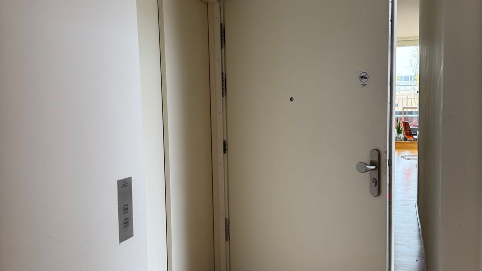 Schmaler, weiß gestrichener Flur mit Holzfußboden und geöffneter weißer Eingangstür mit Türspion und Metallklinke. Links ist eine geschlossene Innentür zu sehen, am Ende des Gangs öffnet sich der Blick in einen hellen Raum mit großen Fenstern.