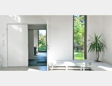 Moderner Innenraum mit weißen Wänden, einem großen Fenster mit Blick auf einen grünen Garten und einem niedrigen weißen Sofa im Vordergrund. Im Hintergrund öffnet sich eine Tür zu einem weiteren Raum mit Zugang nach draußen.