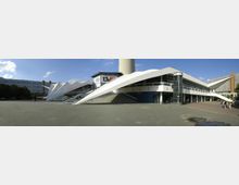 Weitläufiger Platz vor einem modernen Gebäude mit markanter weißer Betonarchitektur, spitz zulaufenden Seitenelementen und einem gläsernen Rundturm. Im Hintergrund ist der Fuß des Berliner Fernsehturms zu erkennen, umgeben von weiteren Gebäuden.