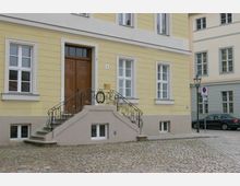 Gelbes mehrstöckiges Gebäude mit weißen Fensterrahmen und einem großen braunen Eingangstor, das über eine Stufenanlage mit verziertem Eisengeländer zugänglich ist; davor ein gepflasterter Platz, links und rechts angrenzende Gebäude sowie eine Straßenlaterne und ein parkendes Auto.