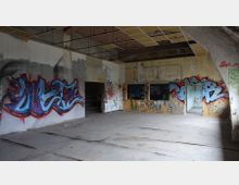 Verlassener Raum mit graffiti-bedeckten Wänden, teilweise zerstörtem Boden und freigelegtem Betongerüst. Die Decke zeigt offene Paneele und herunterhängende Kabel, während kleinere Räume durch offene Türen und Fenster sichtbar sind.