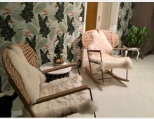 Zwei Schaukelstühle mit geflochtenen Holzlehnen und weißen Fellauflagen stehen in einem Raum mit heller Einrichtung und einer Tapete mit tropischem Pflanzenmotiv. Neben den Stühlen befindet sich ein kleiner Beistelltisch mit einer Lampe und Dekoration.