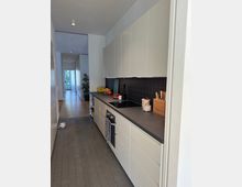 Schmale, moderne Küche mit weißen Schränken, dunkler Arbeitsplatte und schwarzer Fliesenrückwand. Im Hintergrund führt ein langer, heller Flur mit Holzboden zu einer Glastür mit Blick nach draußen.