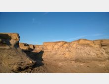 Sandige Felsformationen in einer trockenen, wüstenartigen Landschaft unter klarem, blauem Himmel, mit klar sichtbaren Erosionsspuren an den Geländekanten.