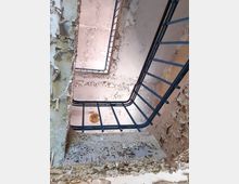 Blick nach oben in einem Treppenhaus eines verlassenen Gebäudes mit abgeblätterter Farbe an den Wänden und blauen Metallgeländern. Eine einzelne Glühbirne hängt von der Decke.