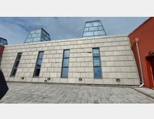 Flaches Gebäude mit einer Fassade aus rechteckigen, hellen Betonplatten, vertikalen schmalen Fenstern und gläsernen gerahmten Pyramiden oberhalb des Daches. Im Vordergrund befindet sich ein gepflasterter Platz, rechts ist ein rotes Wandsegment mit einer Dachrinne sichtbar.