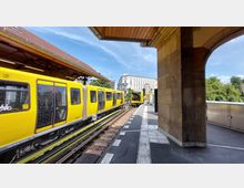 Gelber U-Bahn-Zug an einem erhöhten Bahnhof mit Bahnsteigüberdachung und Steinbögen im Vordergrund; im Hintergrund sind ein weißes historisches Gebäude und grüne Bäume zu sehen.