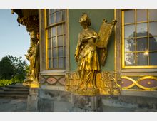 Detailaufnahme eines historischen Gebäudes mit einer vergoldeten Statue einer Frau, die eine Harfe hält, an der Fassade. Die Fassade ist grün mit goldenen Verzierungen und Fenstern mit goldenen Rahmen; im Hintergrund sind Bäume sichtbar.