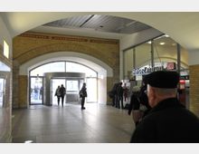 Innenbereich eines Bahnhofs mit gelben Ziegelwänden, einem großen gebogenen Durchgang und einer gläsernen Automatentür im Hintergrund. Rechts befindet sich das „ReiseZentrum“ der Deutschen Bahn mit Schaltern und Automaten, mehrere Personen sind im Raum verteilt.