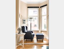 Gemütlicher Erker mit drei großen Fenstern, einem blauen Sessel mit passendem Hocker, einer Stehlampe, einem kleinen weißen Beistelltisch und Parkettboden. Die helle Wandfarbe und die Fenster lassen viel Tageslicht herein.