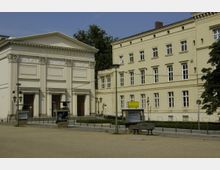 Historisches Gebäudeensemble mit klassizistischer Architektur: Das Maxim Gorki Theater mit seiner symmetrischen Säulenfassade und einem Giebel steht neben einem gelblich-beigen, mehrstöckigen Verwaltungsgebäude. Im Vordergrund sind ein Platz mit einer Bank, Laternen und Infotafeln zu sehen.