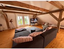 Wohnzimmer mit Holzfußboden und sichtbaren Balken an der Decke. Im Raum befinden sich ein graues Sofa mit gestreiften Kissen, ein Fernseher auf einem Holzmöbel, ein Sessel, ein Teppich sowie eine große Fensterfront mit weißen Vorhängen.