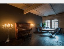 Ein stilvoller Raum mit dunklen Wänden und hohen Decken, ausgestattet mit einem hölzernen Klavier, zwei silbernen Kerzenleuchtern, einer Ledercouch, Sesseln und einem Holztisch. Große Bogenfenster lassen Tageslicht herein, und Pflanzen ergänzen die Einrichtung.