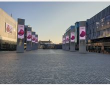 Großer, moderner Platz mit Kopfsteinpflaster, umgeben von Gebäuden mit Glas- und Metallfassaden. Mehrere beleuchtete Werbesäulen mit Anzeigen sind in einer Reihe angeordnet, und im Hintergrund befinden sich ein überdachter Eingang sowie weitere Gebäude.