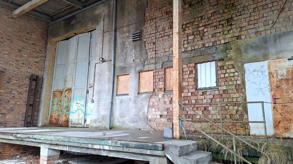 Verwitterte Laderampe eines alten Industriegebäudes mit großer, rostiger Doppeltür und gemauerten Ziegelwänden mit abblätterndem Putz. Mehrere kleine, teils vernagelte Fenster, ein Metallgeländer an einer Betontreppe sowie sichtbare Rohre und Leitungen prägen die Fassade.