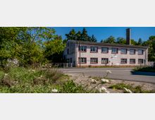 Zweistöckiges, rechteckiges Gebäude mit rosa-verputzter Fassade und mehreren Fenstern, umgeben von Bäumen und Wildwuchs; im Hintergrund ein hoher Kamin und blauer Himmel.