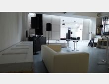 Ein heller, offener Veranstaltungsraum mit weißen Wänden und großen Deckenbeleuchtungen. Im Vordergrund stehen ein helles Sofa und vereinzelte Tische, im Hintergrund sind Lautsprecher, gestapelte Stühle und Veranstaltungstechnik zu sehen.