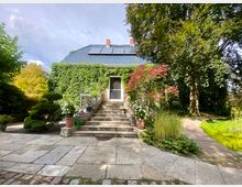 Einfamilienhaus mit begrüntem Dach und einer Fassade, die teilweise von Kletterpflanzen bedeckt ist. Vor dem Eingang führt eine Steintreppe hinauf, umgeben von Topfpflanzen, Bäumen und einem gepflegten Garten mit Holzsteg.