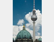 Kuppel des Berliner Doms mit grünlicher Patina im Vordergrund und der Fernsehturm mit kugelförmiger Aussichtsplattform im Hintergrund vor einem Himmel mit weißen Wolken.