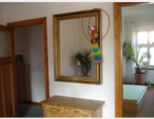 Innenraumansicht mit einem kleinen Holztisch unter einem großen, goldgerahmten Spiegel an einer weißen Wand. Im Spiegel sind eine Zimmerpflanze und ein Fenster mit Tageslicht zu sehen, daneben ist ein dekorativer Papagei an den Rahmen gehängt. Im Hintergrund befindet sich ein Raum mit einem Bett und weiteren Pflanzen.