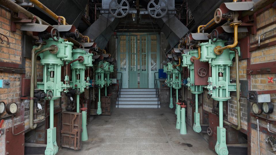 Industrieller Maschinenraum mit historischen hydraulischen Pumpen, die in zwei symmetrischen Reihen an den Wänden montiert sind; im Hintergrund eine Treppe, die zu zweiflügeligen, grünen Glastüren führt. Die Umgebung besteht aus Ziegelwänden und sichtbaren Metallrohren und Hebeln.