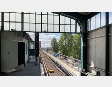 Bahnhof mit Gleisen, die aus einem überdachten Bereich ins Freie führen. Im Vordergrund ist ein Signal zu sehen, im Hintergrund Bäume und ein freier Himmel.