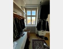 Ein begehbarer Kleiderschrank mit Kleiderstangen und Regalen an beiden Seiten, darauf Kleidung auf Bügeln. In der Mitte steht ein heller Schrank, daneben ein geflochtener Wäschekorb. Das Fenster am Ende des Raums bietet einen Blick auf eine Stadtlandschaft bei klarem Himmel.