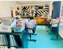 Ein kleiner Schallplattenladen mit türkisfarbenem Boden, hellen Wänden und Regalen voller Vinyl-Platten in der Mitte des Raums. Im Hintergrund befindet sich eine Bar mit Holztheke, dekoriert mit Pflanzen und schwarzen, diagonalen Streifen an der Wand.