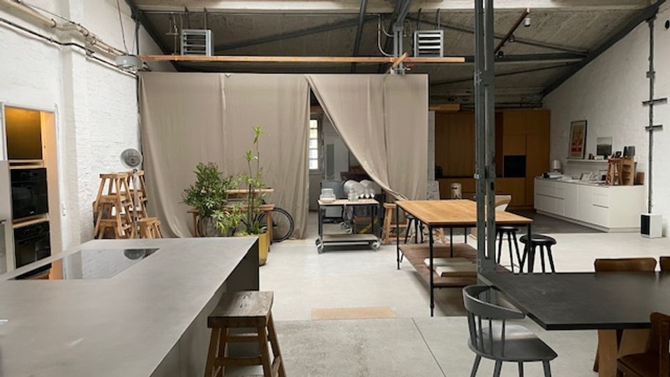 Offener, loftartiger Innenraum mit Betonboden und sichtbaren Deckenleitungen, ausgestattet mit langen Arbeits- und Esstischen, Hockern und einer Küchenzeile an der rechten Wand. In der Mitte trennt ein beiger Vorhangbereich mit Pflanzen und Rollwagen den Raum; links steht eine große Kücheninsel mit Arbeitsfläche.