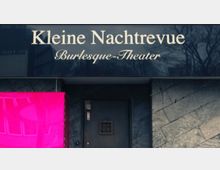 Fassade eines Gebäudes mit der Aufschrift „Kleine Nachtrevue Burlesque-Theater“ auf einem schwarzen Schild. Darunter ein Eingangsbereich mit dunkler Tür und einer hellrosa beleuchteten Fläche links.