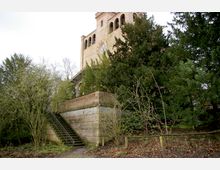 Backsteingebäude mit einem Turm und bogenförmigen Fenstern, umgeben von dichten Bäumen. Eine steinerne Treppe mit Metallgeländer führt von einem Weg hinauf zu einer erhöhten Terrasse.
