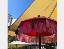 Rosa Sonnenschirm mit Fransen und dekorativen Elementen im Vordergrund, darüber gelb-weiß gestreifte Markise. Im Hintergrund sind ein Gebäude sowie weitere Sonnenschirme in einem Außenbereich sichtbar.