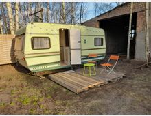 Ein kleiner, hellgelber Wohnwagen der Marke Tabbert steht auf einer Palette in einer ländlichen Umgebung mit Birkenbäumen und einer Backsteinmauer im Hintergrund. Vor dem Wohnwagen befinden sich ein kleiner grüner Tisch und zwei orange Klappstühle.