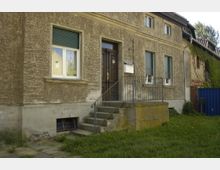 Alt-Text: Außenansicht eines älteren Wohnhauses mit rau verputzter Fassade. Zu sehen ist eine Eingangstür mit einer kleinen Treppe und Metallgeländer, flankiert von Fenstern mit heruntergelassenen Rollläden. Im Vordergrund befindet sich eine Grünfläche, im Hintergrund ist eine mit Efeu bewachsene Wand zu erkennen.