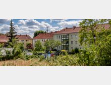 Mehrfamilienhäuser mit pastellfarbenen Fassaden und roten Ziegeldächern liegen inmitten von Bäumen und Grünflächen, im Vordergrund ein Garten mit Wäscheleinen. Der Himmel ist leicht bewölkt, was auf sonniges Wetter hindeutet.