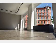 Leerer, moderner Innenraum mit hellen Wänden und großen Glasfenstern, durch die ein historisches, rosa-rot gestrichenes Gebäude im Stadthintergrund sichtbar ist. Der Boden ist aus poliertem Beton, und an der Decke befindet sich eine schwarze Schienenbeleuchtung.