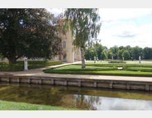 Park mit gepflegten Hecken, Blumenbeeten und Statuen; im Hintergrund ein helles Gebäude mit klassischer Architektur, im Vordergrund ein Wassergraben mit Holzbefestigung.