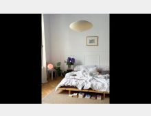 Schlafzimmer mit einem niedrigen Holzbett, bedeckt von unordentlicher weißer Bettwäsche. Im Hintergrund an der weißen Wand befinden sich eine gerahmte Zeichnung, Pflanzen, Bücher und eine Lampe mit warmem Licht, während eine schlichte Papierlampenleuchte von der Decke hängt.