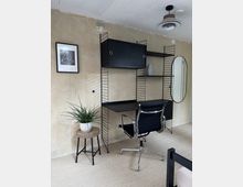 Arbeitszimmer mit schlichtem Design: Ein filigranes schwarzes Regalsystem mit integriertem Schreibtisch an einer unbemalten Wand, daneben ein runder Spiegel. Ein schwarzer Bürostuhl steht vor dem Schreibtisch, und auf einem kleinen Beistelltisch steht eine Zimmerpflanze in einem weißen Topf.