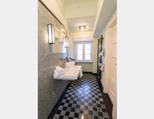 Schmales Badezimmer mit schwarz-weiß kariertem Fliesenboden, grauer Wandverkleidung und langem Spiegel über einem wandmontierten Waschbecken. Am Ende des Raums steht ein Doppelfenster, rechts sind ein schwarzer Handtuchheizkörper und eine weiße Tür zu sehen.