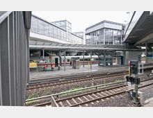 Bahnhofsszene mit mehreren Gleisen im Vordergrund und einem überdachten Bahnsteig im Hintergrund. Auf dem Bahnsteig befindet sich ein ICE-Zug, Sitzgelegenheiten sowie ein Fahrstuhl, umgeben von moderner Glas- und Stahlarchitektur; ein Schild zeigt den Namen "Berlin Südkreuz".