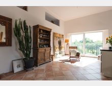 Wohnraum mit heller Einrichtung, großen Fenstern und Terrakottafliesen. Im Vordergrund stehen ein Holzbuffet, ein großer Kaktus in einem Topf und ein Sessel, während durch die Glas-Schiebetüren ein Ausblick ins Grüne sichtbar ist.