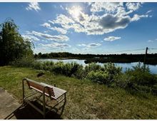 Eine Holzbank steht auf einer Wiese neben einem Flussufer, umgeben von dichter grüner Vegetation. Im Hintergrund ist ein klarer blauer Himmel mit weißen Wolken zu sehen, durch den die Sonne scheint.
