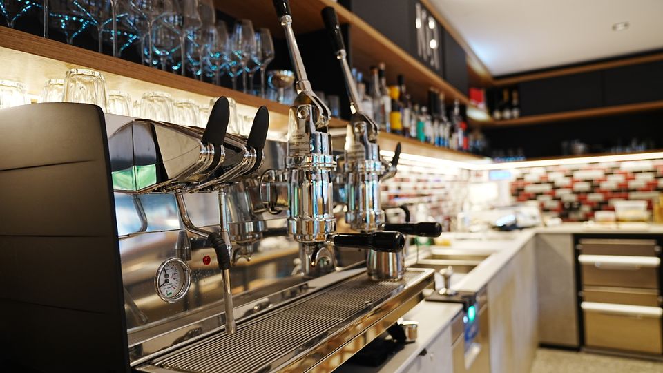 Innenansicht einer modernen Café- oder Bar-Theke mit großer Edelstahl-Espressomaschine im Vordergrund, darüber Regale mit Gläsern und Spirituosenflaschen. Im Hintergrund sind eine beleuchtete Arbeitsfläche, Spüle und eine geflieste Rückwand mit rechteckigem Muster zu sehen.
