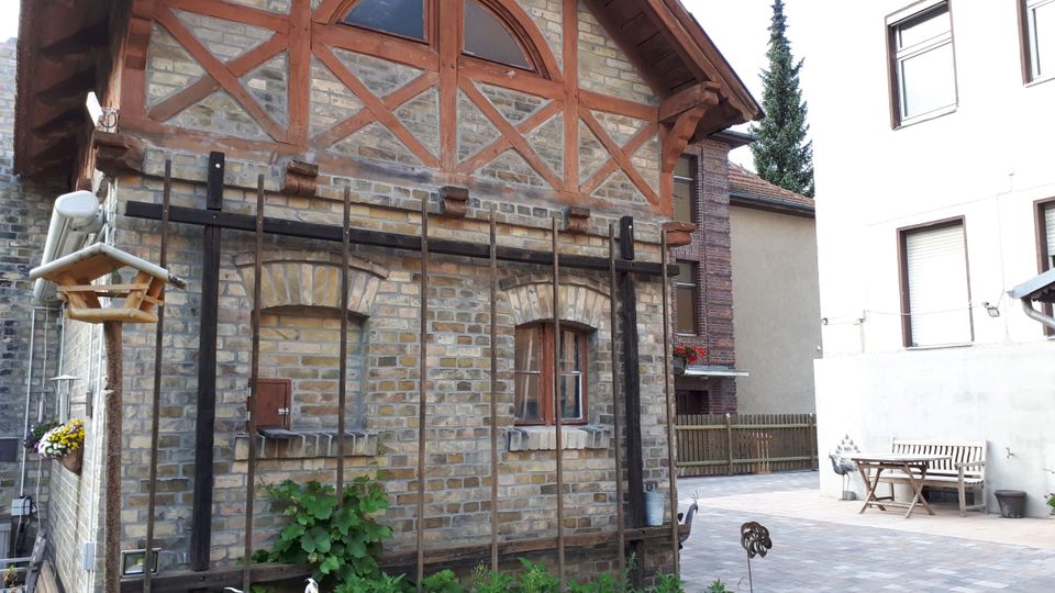 Kleines Backsteingebäude mit Fachwerk-Giebel und halbrundem Fenster im oberen Bereich, davor ein Rankgerüst an der Fassade. Daneben liegt ein gepflasterter Innenhof mit Sitzbank und Tisch zwischen angrenzenden Wohnhäusern.