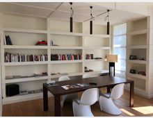 Helles Arbeitszimmer mit weiß gestrichenen Wänden und einer großen, eingebauten Regalwand, die Bücher und Dekorationsgegenstände enthält. Ein dunkler Holztisch mit vier weißen, modern geformten Stühlen steht im Vordergrund, und eine Stehleuchte sowie große Fenster lassen viel Tageslicht herein.