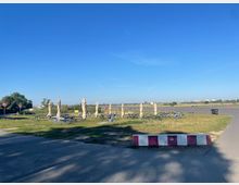 Eine offene Grünfläche mit mehreren gestapelten Sitzgarnituren und geschlossenen Sonnenschirmen, umgeben von Gehwegen und einem asphaltierten Platz. Im Hintergrund sind eine weite Landschaft und einige Gebäude am Horizont zu sehen, unter klarem, blauem Himmel.