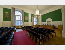 Innenansicht eines historischen Sitzungssaals mit hohen, grünen Wänden, großen Fenstern mit Rundbögen und Vorhängen. Der Raum ist mit einer hölzernen Bühne, schwarzen Stühlen in Reihen, einem großen Kronleuchter und Wandverkleidungen in Weiß dekoriert.